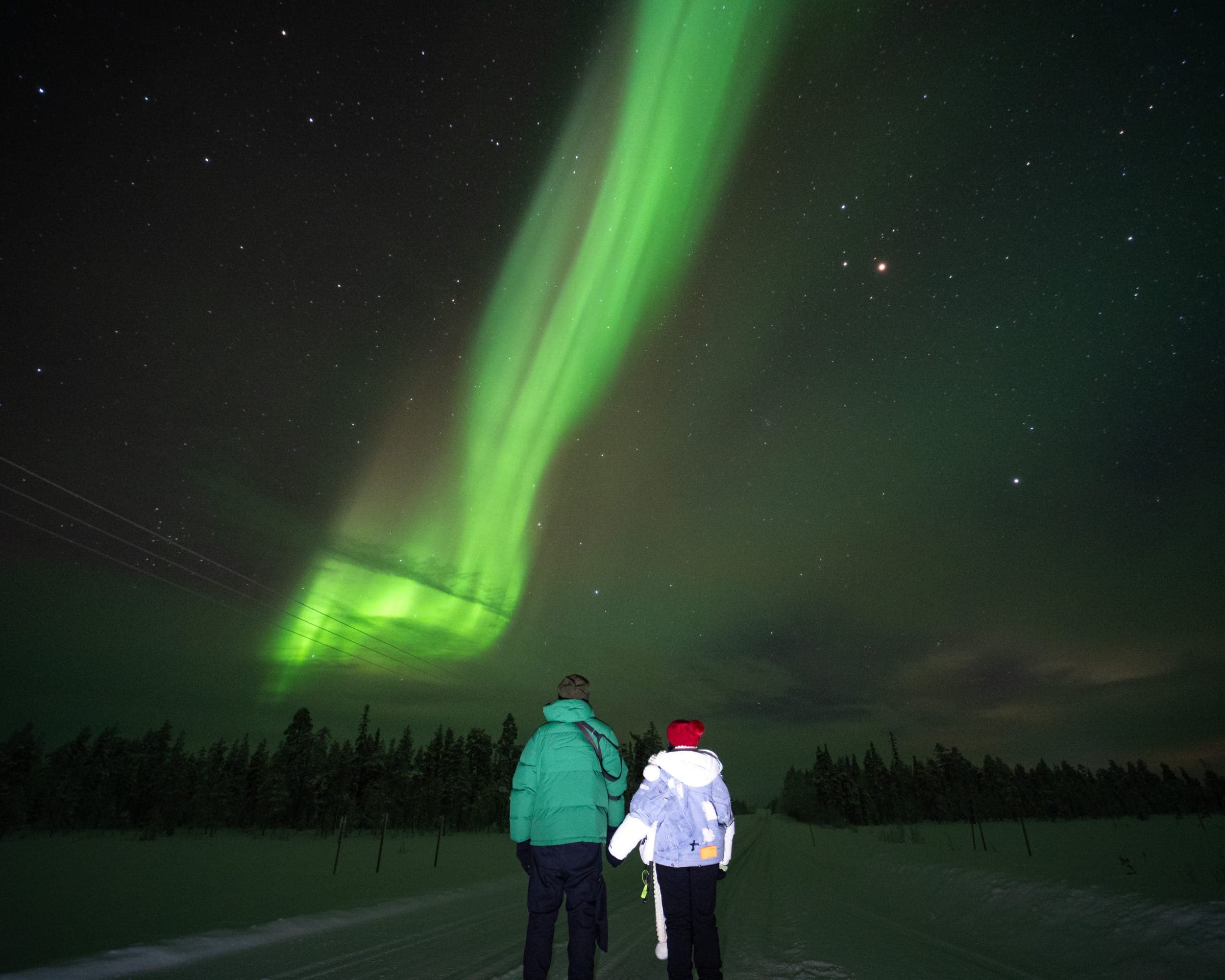 Two guests on a snowy road beneath a tall arc of northern lights